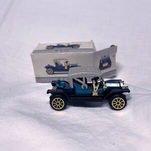 Può includere: Un'auto giocattolo blu e nera con un tetto grigio e ruote dorate. L'auto è un modello vintage e nello stile di un'auto degli anni '20. L'auto è seduta su una superficie bianca.