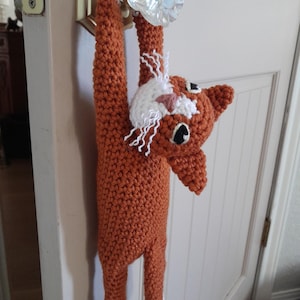 Puede incluir: Un gato de crochet naranja y blanco colgado de un pomo de puerta. El gato tiene una cara blanca con ojos negros y una nariz rosa.