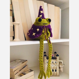 May include: A handmade crochet frog wearing a purple wizard hat and cape with gold stars. The frog is green with black eyes and long, dangling green legs. The frog is sitting on a white shelf with books in the background.