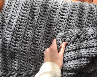 May include: A gray crocheted blanket with a cable knit pattern. The blanket is folded in half and a hand is reaching out to touch the fabric.