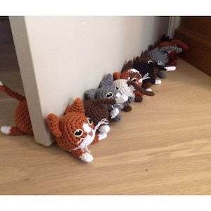 May include: A row of crocheted cat doorstops in various colours, including grey, brown, white, and orange, with white faces and black eyes. The cat doorstops are lined up on a wooden floor.
