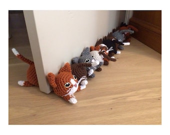 May include: A row of crocheted cat doorstops in various colours, including grey, brown, white, and orange, with white faces and black eyes. The cat doorstops are lined up on a wooden floor.