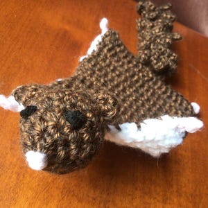 May include: A brown and white crocheted squirrel with black eyes. The squirrel has a long tail and is lying on its side.