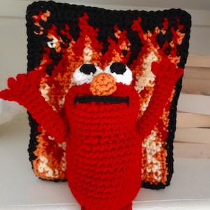 Puede incluir: Una figura de ganchillo roja con una nariz naranja redonda, ojos negros y boca negra, colocada frente a un panel rectangular de ganchillo con un patrón de llamas en tonos negro, naranja y blanco con ambos brazos levantados.