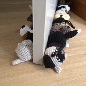 May include: A group of crocheted dog toys in various colors and patterns, including black, white, brown, and tan. The toys are arranged in a line, with their heads facing the viewer. They are placed against a white door frame.