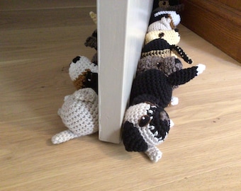 May include: A collection of crocheted dog toys in a variety of colours and patterns, including black, white, brown, and tan. The toys are lined up, with their heads facing the viewer. They are positioned against a white door frame.