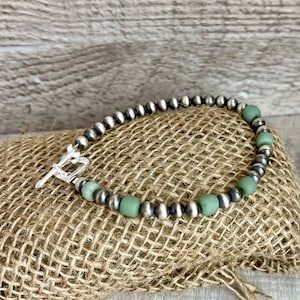 May include: A silver and green beaded bracelet with a silver clasp. The bracelet is on a burlap background.