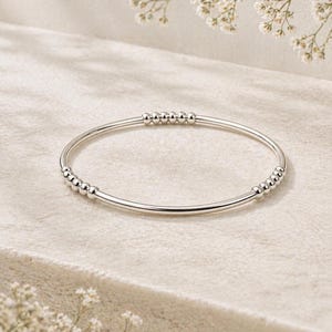 May include: A polished silver-tone bangle bracelet featuring a smooth, thin circular band accented with two distinct groupings of small, round metallic beads positioned along the circumference of the jewelry piece.
