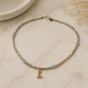 May include: A beaded necklace featuring small, round, metallic gold-tone beads interspersed with translucent, pale blue, faceted gemstone beads. A single, polished, gold-tone letter E charm hangs from the center of the circular necklace strand.