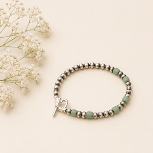 May include: A silver beaded bracelet with a toggle clasp. The bracelet features small, round silver beads interspersed with square, light green beads. The bracelet is displayed on a cream-colored surface, with white flowers in the background.
