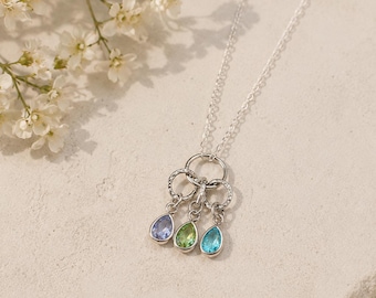 May include: A silver necklace with three teardrop-shaped pendants in blue, green, and turquoise. The pendants are suspended from a cluster of textured rings. The necklace is displayed on a light-colored surface with white flowers in the background.