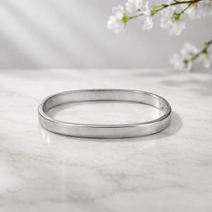 May include: A polished silver oval bangle bracelet. The bracelet has a smooth, flat surface and a simple, elegant design. The background is a light marble surface with white flowers.