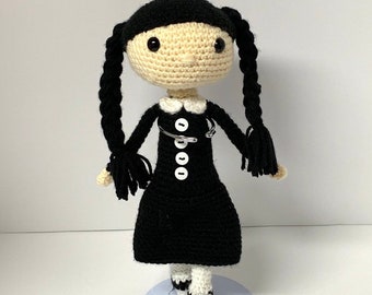 May include: A crocheted doll with black hair in pigtails, wearing a black dress with white buttons and a safety pin, and white and black striped socks.