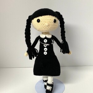 Puede incluir: Una muñeca de crochet con el pelo negro en trenzas, que lleva un vestido negro con botones blancos y un imperdible, y calcetines a rayas blancas y negras.