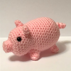 May include: A handmade, light pink crochet pig with black button eyes, small ears, and a curly tail. The pig has four small legs and a rounded body. This soft toy is a charming addition to any collection.