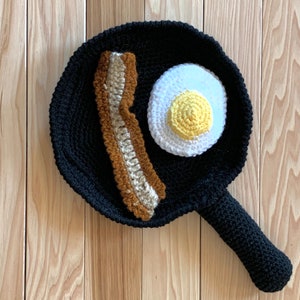 May include: A crocheted black frying pan with a brown and beige crocheted bacon strip and a white crocheted egg with a yellow yolk.