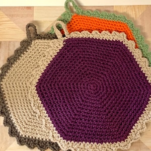 Könnte beinhalten: Drei gehäkelte Topflappen in verschiedenen Farben. Der größte Topflappen ist lila mit einem beigefarbenen Rand und einem braunen Außenrand. Der mittlere Topflappen ist orange mit einem grünen Außenrand. Der kleinste Topflappen ist beige mit einem grünen Außenrand.