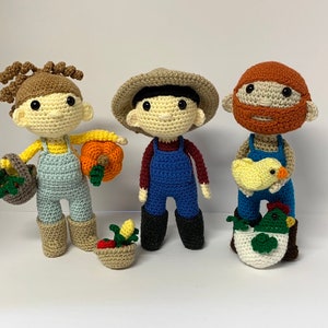 May include: Three crocheted dolls dressed as farmers. The doll on the left is wearing a blue and yellow outfit and holding a pumpkin. The doll in the middle is wearing a blue and red outfit and a brown hat. The doll on the right is wearing a blue and yellow outfit and holding a yellow chick.