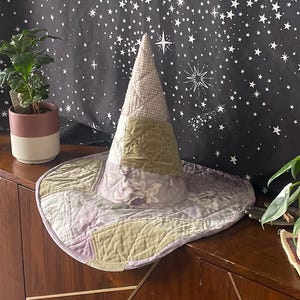 May include: A quilted witch hat with a wide brim and a pointed crown, in shades of lavender, green, and cream. The hat sits on a wooden cabinet with gold accents, in front of a black tapestry with a white star and moon pattern.