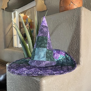 May include: A quilted witch hat featuring a patchwork design in various shades of purple, dark green, and teal. The fabric displays assorted floral and geometric patterns, with visible stitching throughout the conical crown and wide, circular brim.