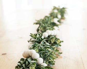 May include: A green garland with white felt balls, perfect for decorating a table for a holiday or special occasion.