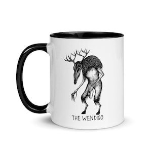 May include: White ceramic coffee mug with a black rim and handle. The mug features a black and white illustration of a creature with antlers and the text "THE WENDIGO".