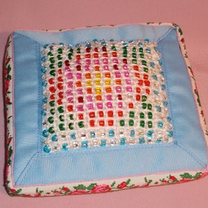 May include: A square blue and white pincushion with a floral design on the border. The center of the pincushion is a colorful beaded design with a rainbow of colors.