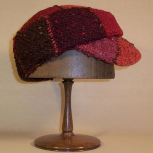 May include: A red, brown and black knitted hat with a patchwork design. The hat is displayed on a wooden hat stand.