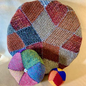 May include: A collection of colorful, handmade fabric items. The image features a large, round patchwork pillow in shades of brown, red, and gray. Two smaller, multi-colored, spherical objects and a smaller ball are also present, showcasing a variety of vibrant hues.
