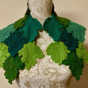 Puede incluir: Un chal hecho a mano con forma de hojas, en varios tonos de verde, expuesto sobre un maniquí. El chal presenta un diseño texturizado y tejido, con cada hoja meticulosamente elaborada. La paleta de colores varía de verde claro a oscuro, creando una estética natural.