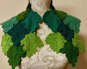 May include: A handmade leaf-shaped shawl in various shades of green, displayed on a mannequin. The shawl features a textured, woven design, with each leaf meticulously crafted. The color palette ranges from light to dark green, creating a natural, organic aesthetic.