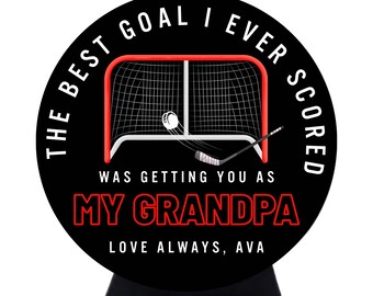 May include: A circular black sign featuring a white hockey goal, a hockey stick, and a puck. The text reads: THE BEST GOAL I EVER SCORED WAS GETTING YOU AS MY GRANDPA, LOVE ALWAYS, AVA. The words MY GRANDPA are in red, while the rest is white.
