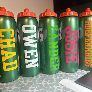 Pode incluir: Cinco garrafas de &aacute;gua desportivas de pl&aacute;stico verde com tampas de rosca cor de laranja. Cada garrafa apresenta um log&oacute;tipo Gatorade e um nome personalizado em letras grandes: CHAD em amarelo, OWEN em branco, ZANDER em verde lima, JOSIE em rosa e HOEFENKRIEG em laranja.