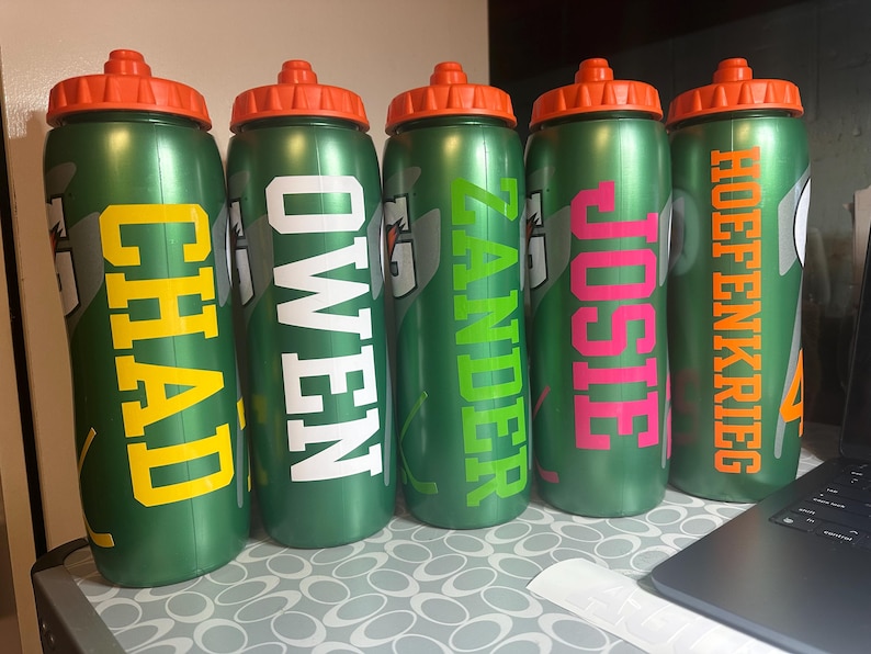 Pode incluir: Cinco garrafas de &aacute;gua desportivas de pl&aacute;stico verde com tampas de rosca cor de laranja. Cada garrafa apresenta um log&oacute;tipo Gatorade e um nome personalizado em letras grandes: CHAD em amarelo, OWEN em branco, ZANDER em verde lima, JOSIE em rosa e HOEFENKRIEG em laranja.
