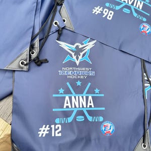 May include: Navy blue drawstring bags featuring the Northwest Icehawks Hockey logo, a graphic of crossed hockey sticks with a puck, and personalized text including the names Anna and Gavin, along with jersey numbers 12 and 98 in white print.