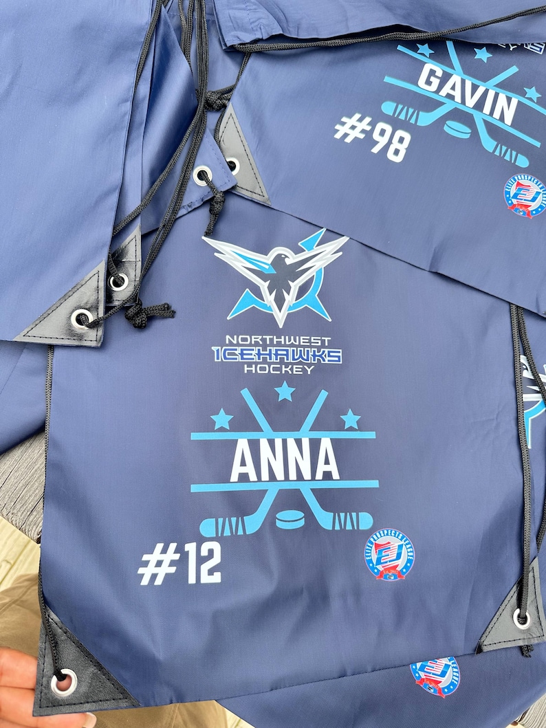 May include: Navy blue drawstring bags featuring the Northwest Icehawks Hockey logo, a graphic of crossed hockey sticks with a puck, and personalized text including the names Anna and Gavin, along with jersey numbers 12 and 98 in white print.