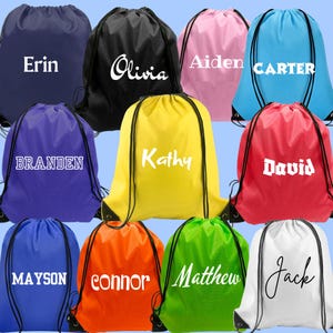 May include: A collection of eleven drawstring backpacks in various solid colors including navy, black, pink, light blue, purple, yellow, red, royal blue, orange, green, and white, each personalized with a unique white name printed on the front.