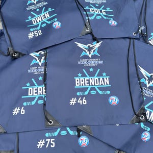 Può includere: Una collezione di sacche con coulisse blu navy con il logo Northwest Icehawks Hockey in bianco e azzurro. Ogni borsa è personalizzata con il nome di un giocatore, un numero di maglia e una grafica di due bastoni da hockey incrociati.
