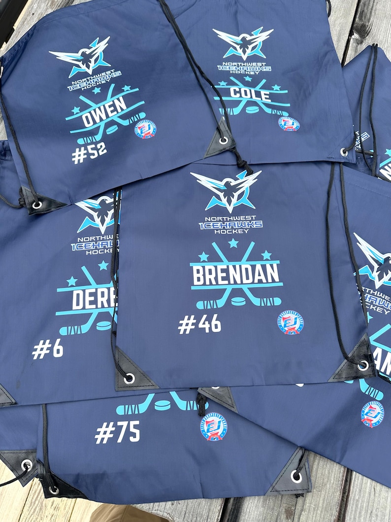 May include: A collection of navy blue drawstring bags featuring the Northwest Icehawks Hockey logo in white and light blue. Each bag is personalized with a player name, a jersey number, and a graphic of two crossed hockey sticks and a puck.