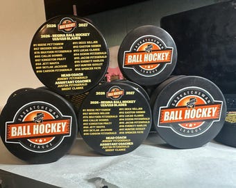 May include: A collection of black hockey pucks featuring the Saskatchewan Ball Hockey League logo in orange and white, alongside custom printed pucks listing the 2026-2027 Regina Ball Hockey U15/U18 Blades team roster, including player names and coaching staff.