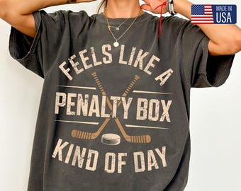 May include: Dark gray t-shirt with the text "FEELS LIKE A PENALTY BOX KIND OF DAY" in a distressed font. The design includes crossed hockey sticks and a puck. Made in the USA.