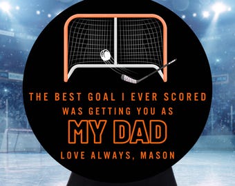 May include: A circular black sign featuring an orange hockey goal graphic with a puck and stick. The text reads: THE BEST GOAL I EVER SCORED WAS GETTING YOU AS MY DAD. LOVE ALWAYS, MASON. The design is set against a blurred hockey rink background.