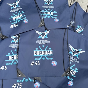 May include: A collection of dark blue drawstring bags featuring the Northwest Icehawks Hockey logo in light blue and white. Each bag is personalized with a player's name, a jersey number, and a graphic of crossed hockey sticks and a puck.