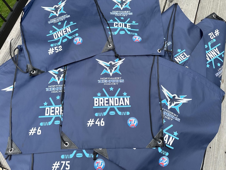 May include: A collection of dark blue drawstring bags featuring the Northwest Icehawks Hockey logo in light blue and white. Each bag is personalized with a player's name, a jersey number, and a graphic of crossed hockey sticks and a puck.