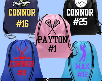 May include: Five drawstring bags in navy, black, pink, blue, and light blue. The navy bag features a pickleball graphic, the name CONNOR, and #16. The black bag shows a volleyball, CONNOR, and #25. The pink bag displays lacrosse sticks, PAYTON, and #1.