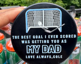 May include: A round black hockey puck featuring a white and light blue graphic of a hockey net and stick. The text reads: THE BEST GOAL I EVER SCORED WAS GETTING YOU AS MY DAD LOVE ALWAYS, COLE. The design is printed in white and light blue.