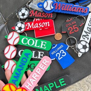 Könnte beinhalten: Eine Sammlung personalisierter Schlüsselanhänger in verschiedenen Farben. Schlüsselanhänger mit Namen wie "Mason", "Cole" und "Amanda" sowie Sportmotiven wie Basebällen, Fußbällen und Trikots. Die Schlüsselanhänger sind aus Kunststoff und haben Metallringe.