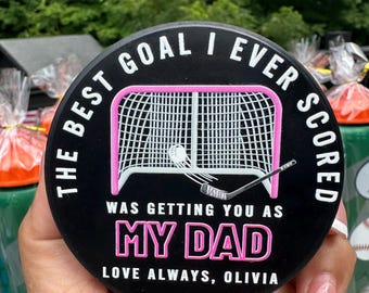 May include: A black circular hockey puck featuring a white graphic of a hockey net and stick. The text reads: THE BEST GOAL I EVER SCORED WAS GETTING YOU AS MY DAD. LOVE ALWAYS, OLIVIA. The words MY DAD are in pink, and the net frame is outlined in pink.