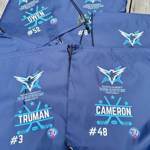 May include: A collection of navy blue drawstring bags featuring the Northwest Icehawks Hockey logo in light blue and white. Each bag is personalized with a player's name, such as Owen, Truman, or Cameron, along with their specific jersey number.