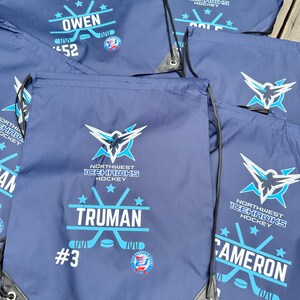 May include: A collection of navy blue drawstring bags featuring the Northwest Icehawks Hockey logo in white and light blue. Each bag is personalized with a player's name, such as Truman, Owen, or Cameron, along with a jersey number and a hockey graphic.